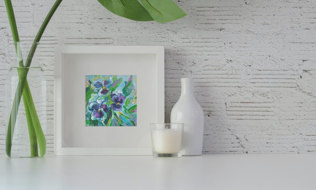 A painting by Mary Haapala of three pansies framed in white, sits on a ledge next to some objects