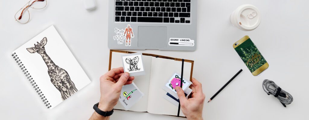 mary haapalas digital designs can be seen on stickers, laptops and phone cases scattered on a desk.