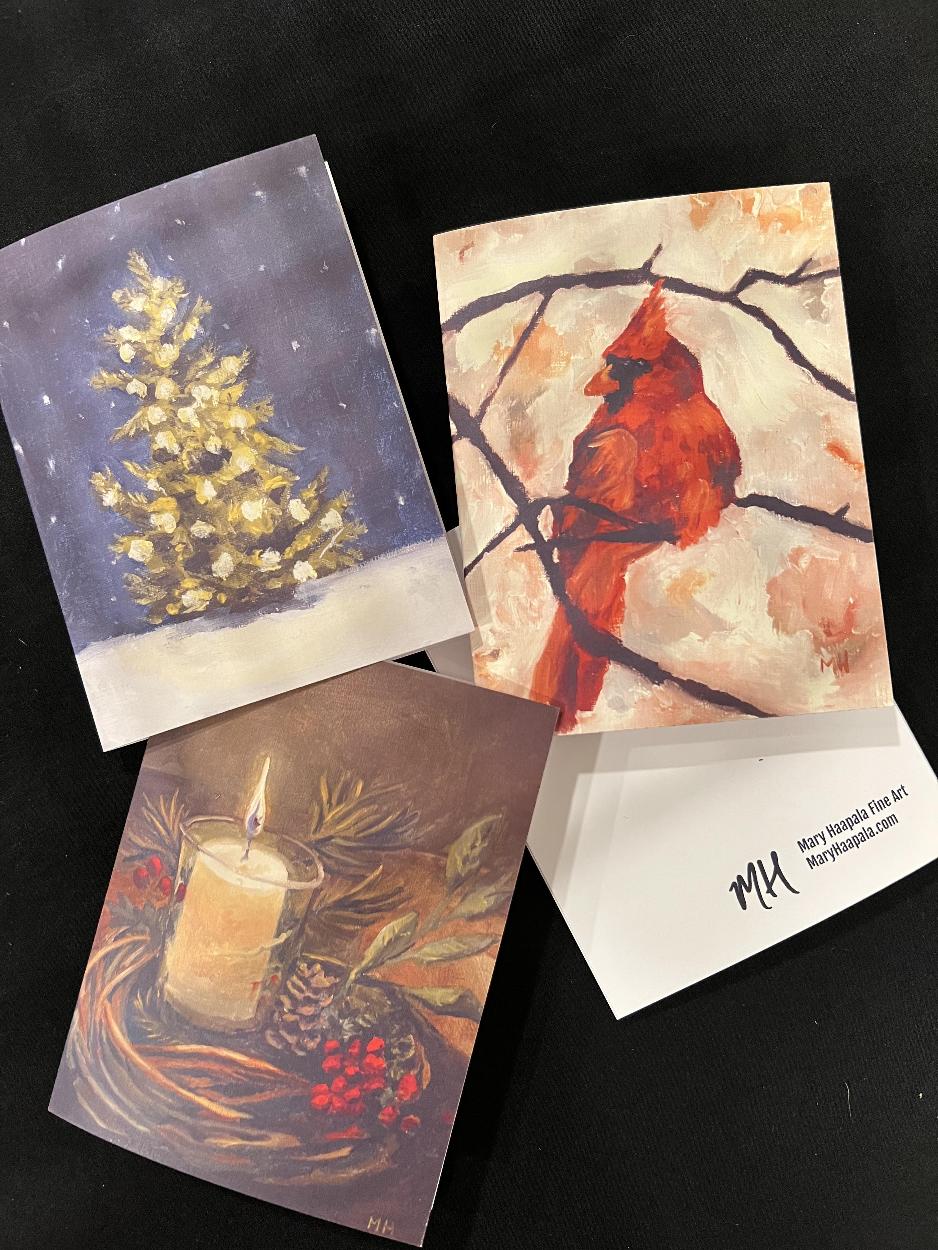 set of 3 greeting cards by mary haapala laying on a table. Cards portray three different designs: christmas tree with lights on a blue background. red cardinal in branches and a lit candle in a twig wreath with red berries and greenery.