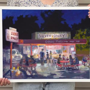 Artist holding framed reproduction of original artwork titled "Silver Dairy". It is a nostalgic piece of art with a view of a local ice cream shop with groups of people enjoying a summer night.