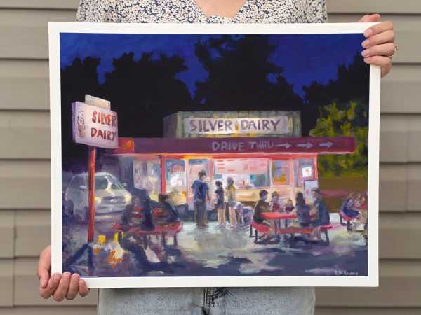 Artist holding framed reproduction of original artwork titled "Silver Dairy". It is a nostalgic piece of art with a view of a local ice cream shop with groups of people enjoying a summer night.