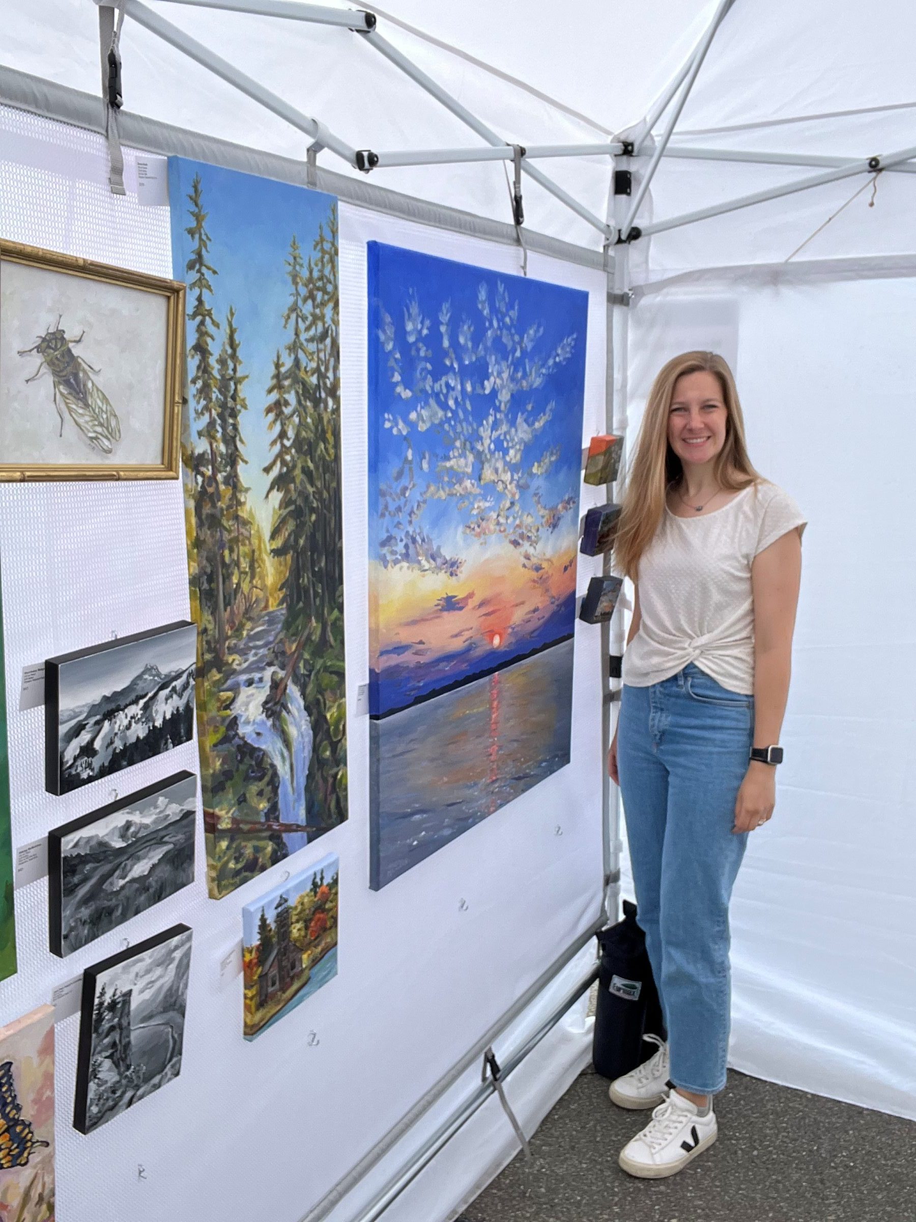 Mary Haapala standing next to some colorful paintings on the wall. One painting is of a sunset, the other a waterfall.